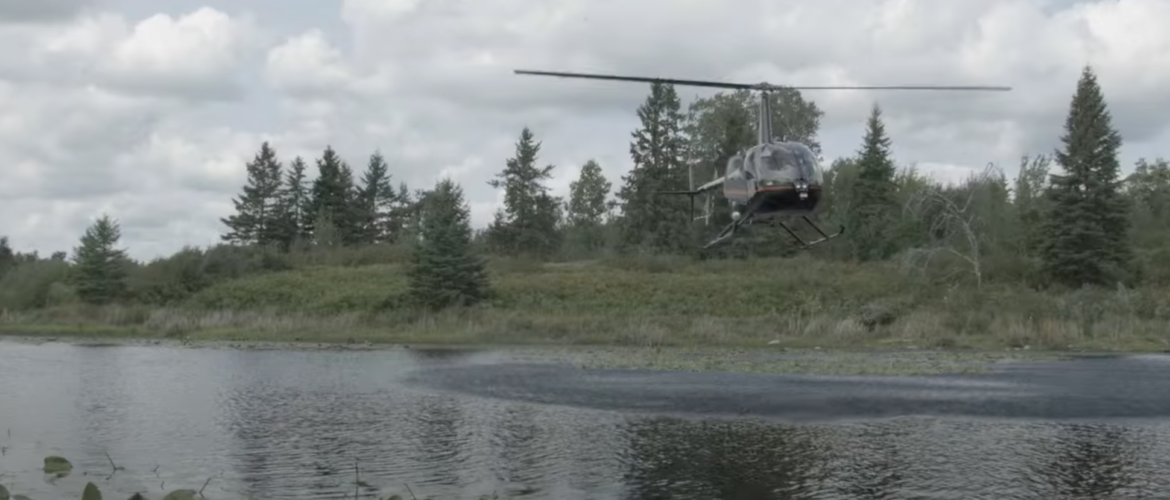 Image shows a helicopter flying over a shallow lake with green forested landscape in the background